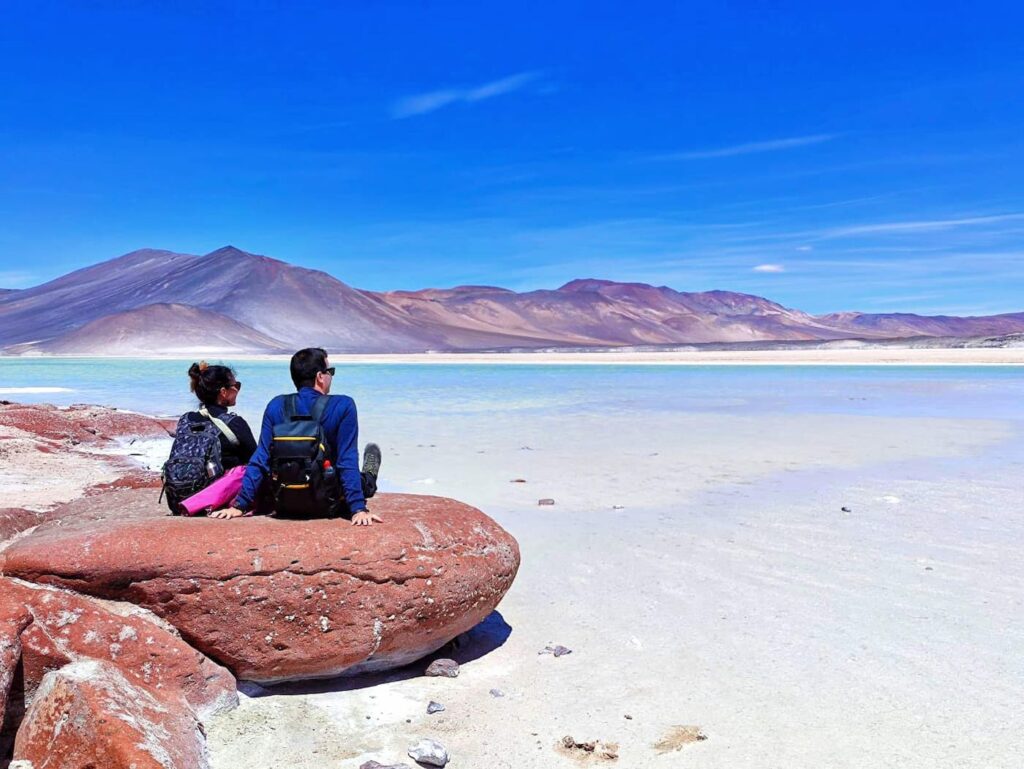 Domi y Montse, de espaldas y a la izquierda de la imagen, contemplan el imponente paisaje del Salar de Piedras Rojas, en el altiplano chileno, con sus formaciones rojizas, lagunas y cielo despejado en una fotografía espectacular de Destino y Maleta.