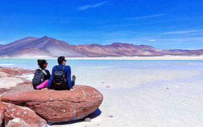 Domi y Montse, de espaldas y a la izquierda de la imagen, contemplan el imponente paisaje del Salar de Piedras Rojas, en el altiplano chileno, con sus formaciones rojizas, lagunas y cielo despejado en una fotografía espectacular de Destino y Maleta.